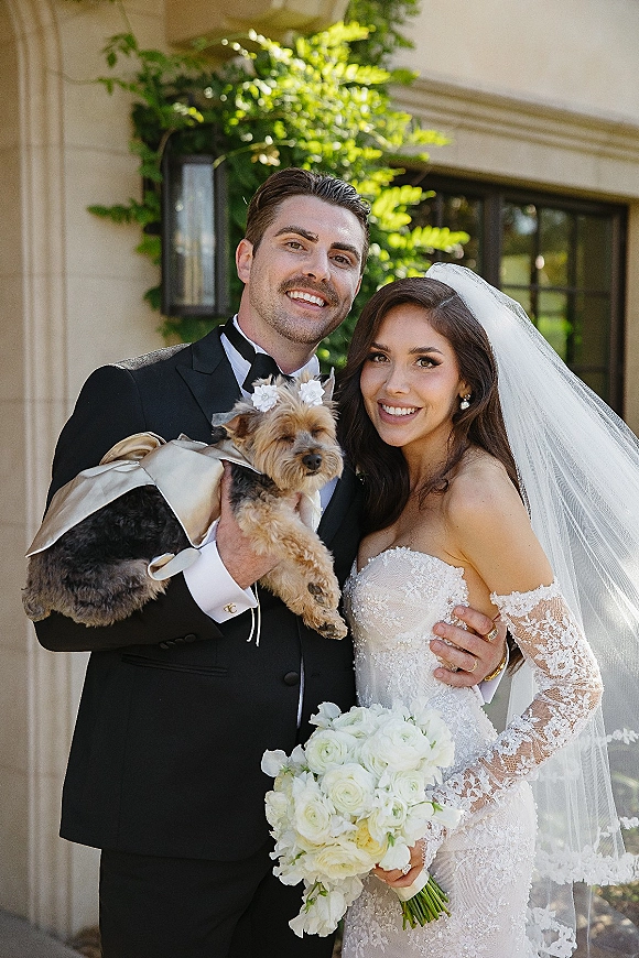 Couple portrait of bride and groom with dog, bride in strapless lace gown and veil holding white rose bouquet outside ivy-covered wall