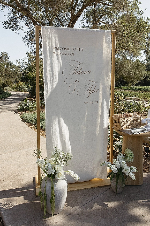 Wedding welcome sign on linen fabric in a wooden frame with calligraphy and white roses, set beside escort cards on a garden walkway