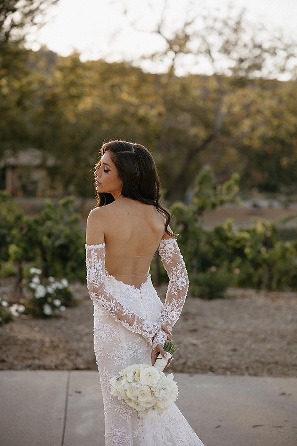 Bridal portrait of a bride in a backless wedding dress holding a white rose bouquet behind her back on a garden walkway