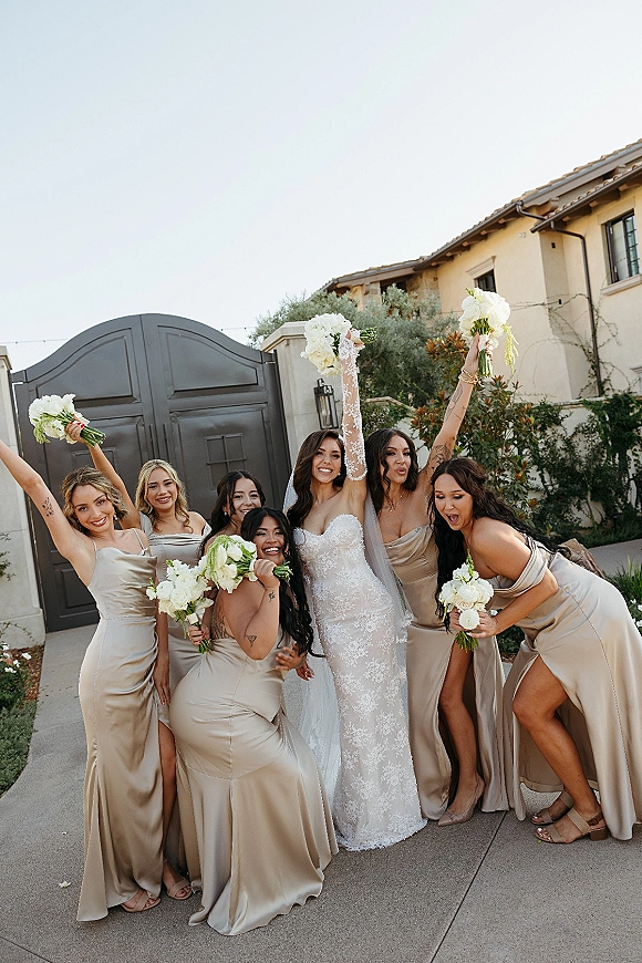 Bridesmaid group photo with bride and bridesmaids in champagne bridesmaid dresses raising white and green bouquets by a villa gate