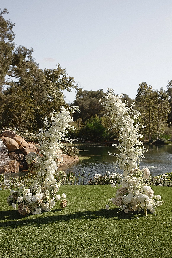 Wedding ceremony arch with asymmetrical floral arch of white roses and hydrangeas, set on a lakeside lawn with rocks and trees behind