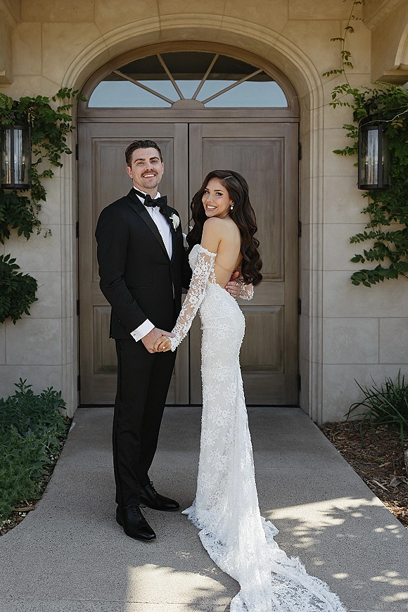 Couple portrait of bride and groom holding hands, full length, by an arched wooden doorway with ivy; she wears a lace train, he a black tuxedo.