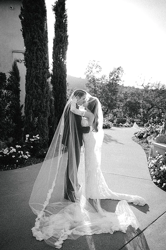 Wedding kiss portrait of bride and groom kissing under a lace-trimmed veil on a garden path lined with tall cypress trees