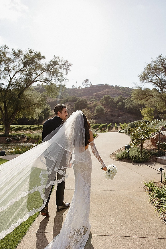 Couple portrait of bride and groom walking away, her cathedral veil trailing, holding a white rose bouquet on a sunlit vineyard path