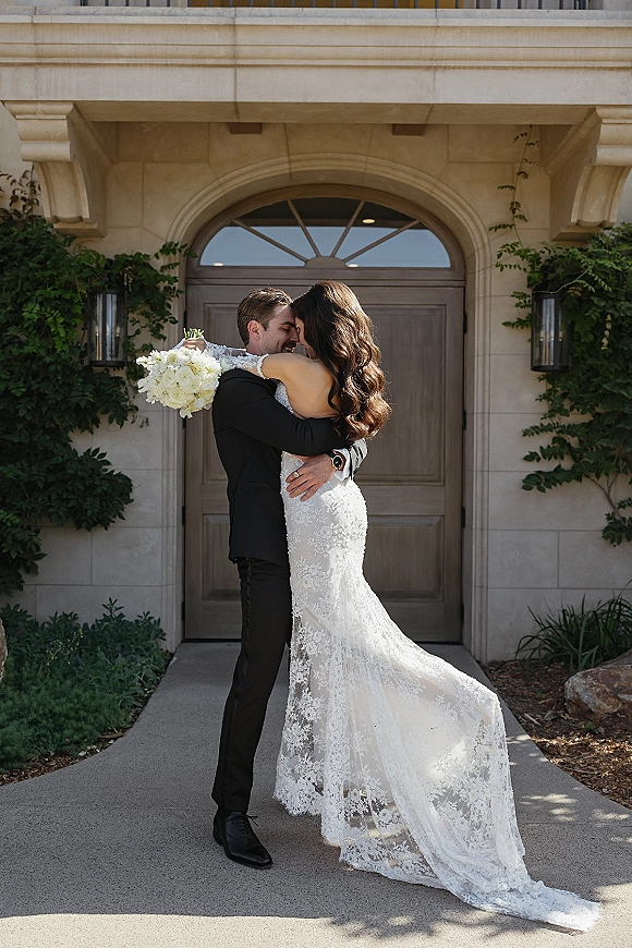 Couple portrait of bride and groom embrace with forehead touch, lace dress train and white bouquet at ivy-framed stone doorway entrance