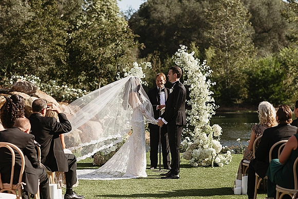 Ceremony moment as bride and groom hold hands at a lakeside outdoor wedding ceremony beneath a floral arch, veil flowing in breeze