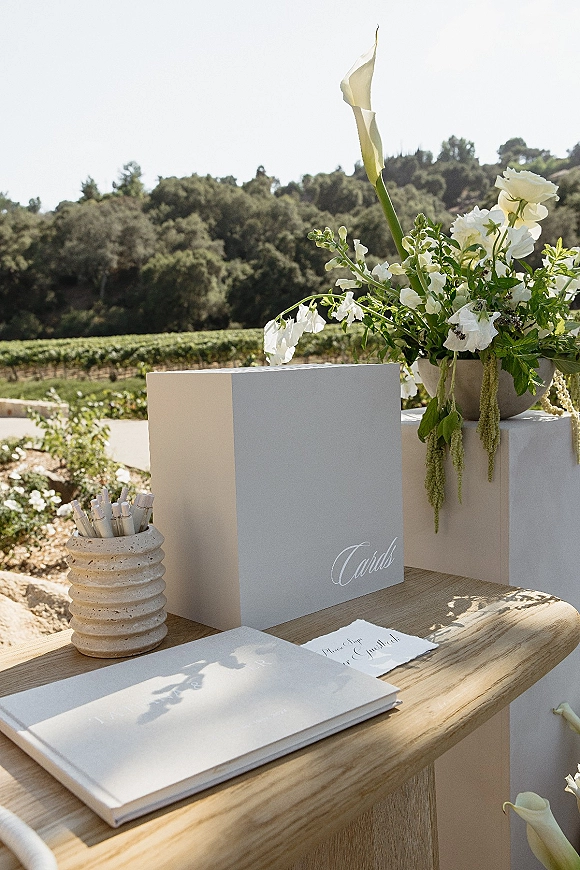 Wedding guest book on a wedding guest book table with pens, card box, and calla lilies on pedestal stands in an outdoor vineyard setting