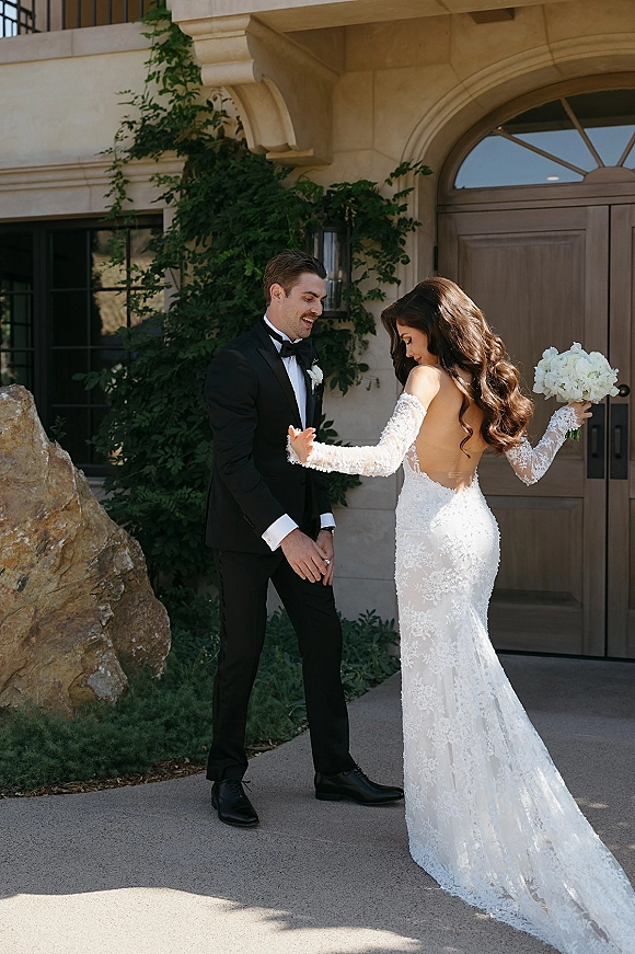 Couple portrait of bride and groom laughing as she turns in a long-sleeve lace gown, holding white bouquet by an ivy stone doorway