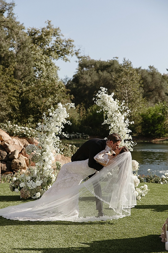 Wedding kiss portrait of bride and groom in a dip kiss wedding photo under a white floral arch by a lakeside lawn with veil flowing