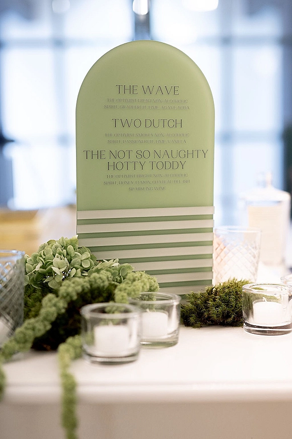 Signature drink menu displayed on an arched wedding bar menu sign with greenery garland, votive candles, and textured glassware by bright windows