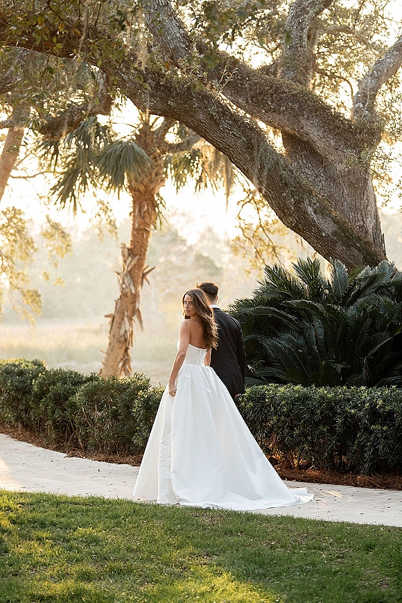 Couple portrait in a garden, bride and groom from behind walking on a brick path as she looks over her shoulder, long train in sunlight