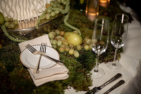 Reception tablescape with a moss table runner, white and gold place settings, candles, and a wedding cake with grapes and pears under indoor lighting