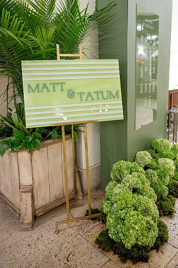 Wedding welcome sign on easel with striped board, hydrangea blooms and greenery, potted palm and lantern on stone tile floor backdrop