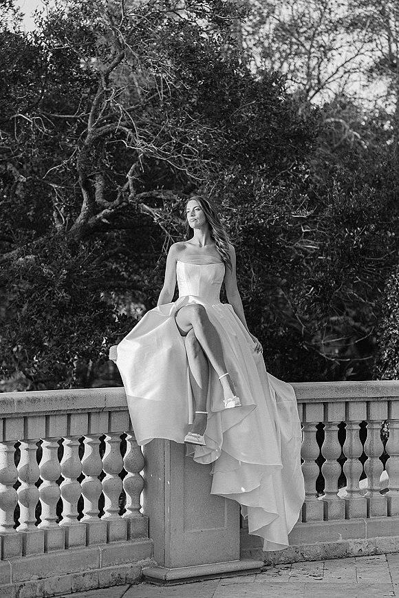 Bridal portrait of a bride in a strapless wedding dress with a high-low skirt, sitting on a stone balustrade by garden trees