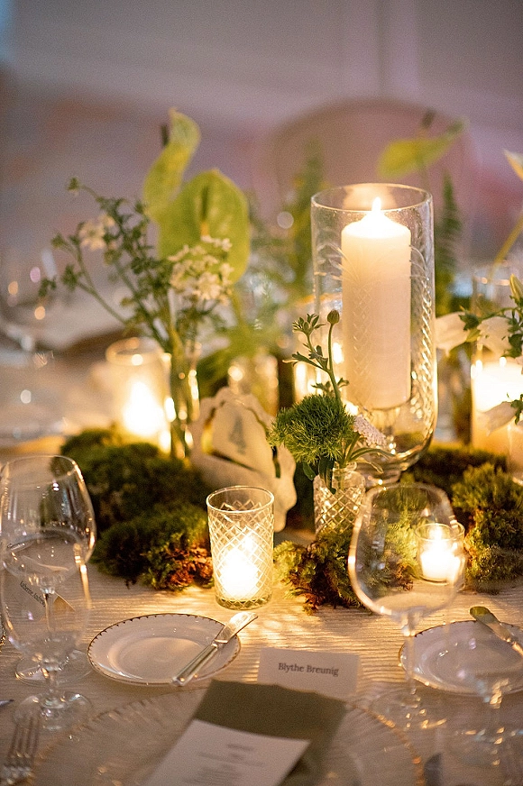 Reception tablescape with a wedding table centerpiece of glass hurricane pillar candles, moss runner, bud vases, and place cards in soft candlelight