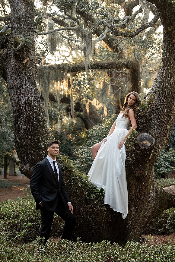 Couple portrait of bride and groom outdoors, bride sitting on an oak tree in a strapless satin dress as he leans in, bouquet greenery in hand