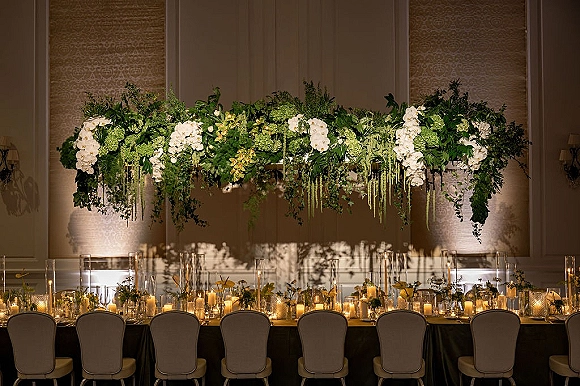 Head table decor with wedding sweetheart table flowers, white orchid and greenery garland, and glowing pillar candles against ballroom wall panels