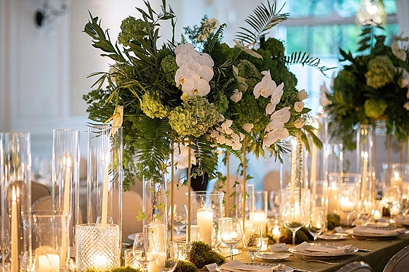 Reception tablescape with wedding table centerpiece of tall white orchid and hydrangea florals, greenery, and floating candles in glass vases near windows
