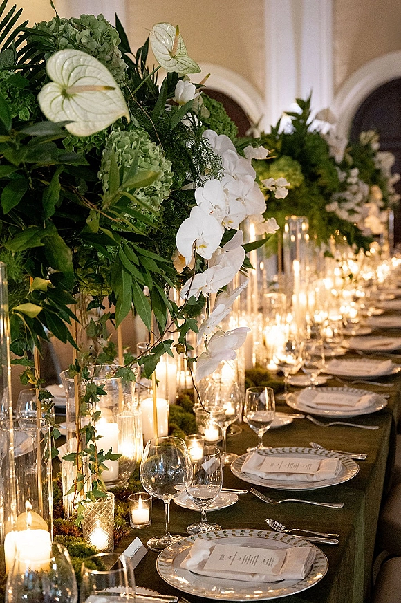 Reception tablescape with green and white tablescape florals, orchids and hydrangea, candlelit hurricane vases, and patterned place settings on a banquet table