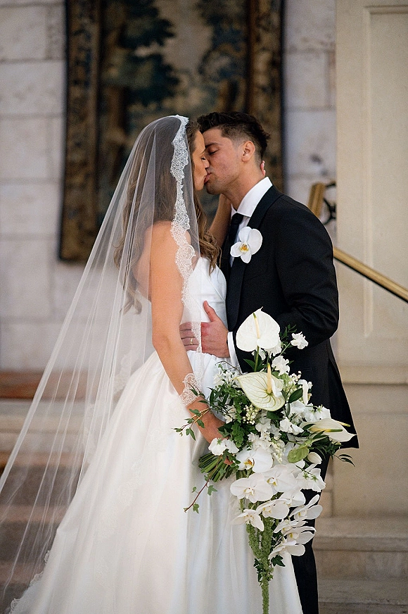 Wedding kiss portrait of bride and groom kissing, her lace veil and cascading orchid bouquet, in a stone interior by a staircase
