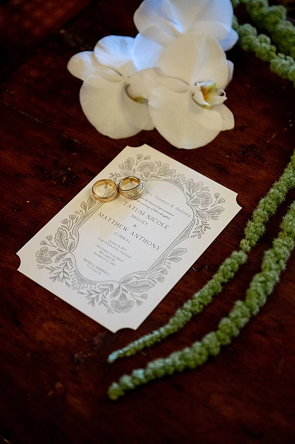 Wedding invitation suite with wedding rings on invitation, engraved floral card and a white orchid on a dark wooden table flat lay