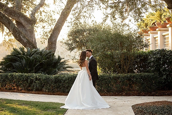 Wedding kiss portrait of bride and groom kissing, holding hands on a stone walkway in a villa garden, golden hour light, strapless gown train