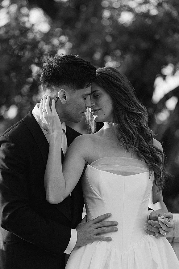 Wedding couple portrait in black and white, bride and groom embrace with foreheads touching as she shows her wedding ring in sunlit garden bokeh