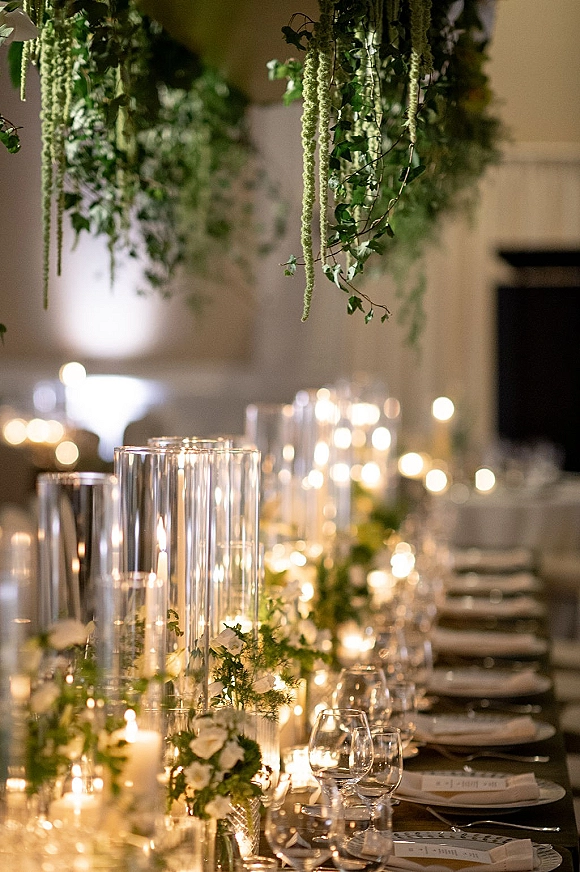 Reception tablescape with candlelit wedding reception table featuring taper candles in glass cylinders, greenery garland, and white flowers under hanging greenery