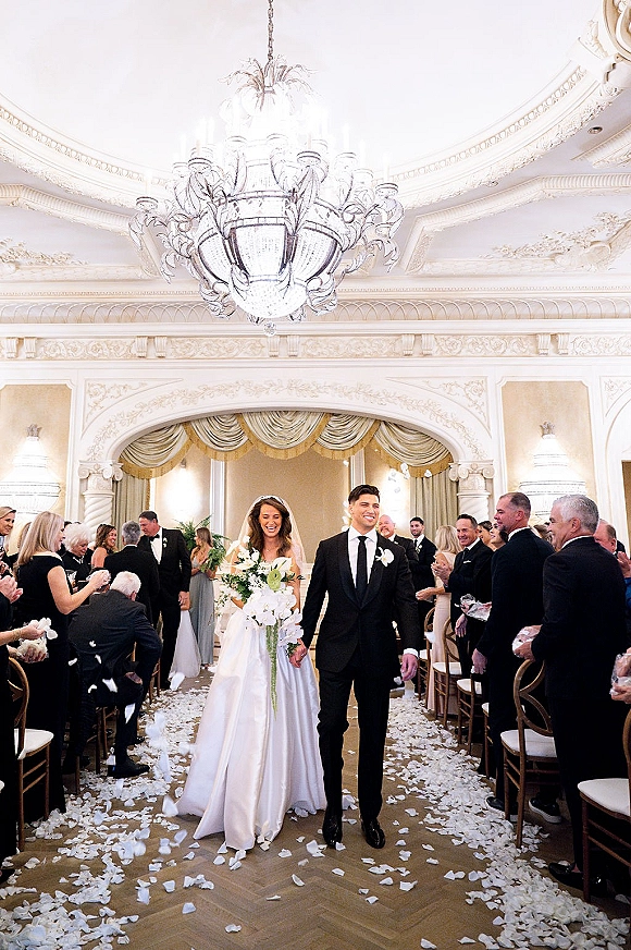 Wedding recessional as bride and groom walk the aisle, she holds a cascading bouquet and veil trails under a grand chandelier in ballroom