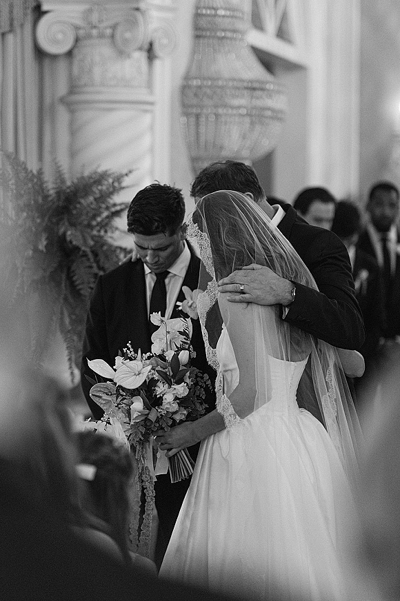 Ceremony moment as bride and groom praying in a church wedding ceremony, groom embracing her under a chandelier, lace veil and bouquet visible