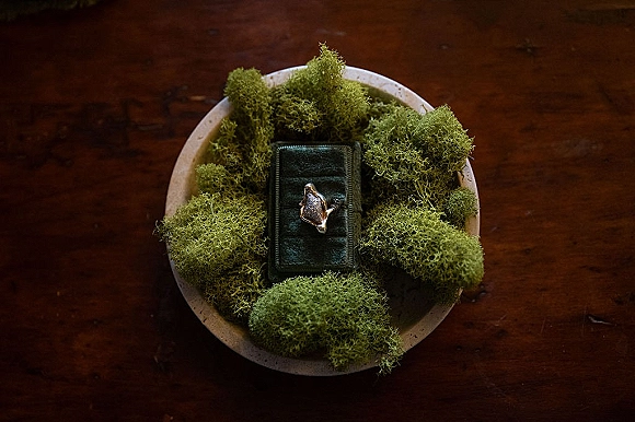 Engagement ring close up resting on moss in a ring box on a ceramic dish, styled on a rustic wood table flat lay