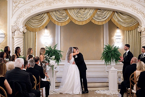 Ceremony kiss as bride in veil and groom in black tuxedo share a kiss at an ornate ballroom altar with arch and draped backdrop