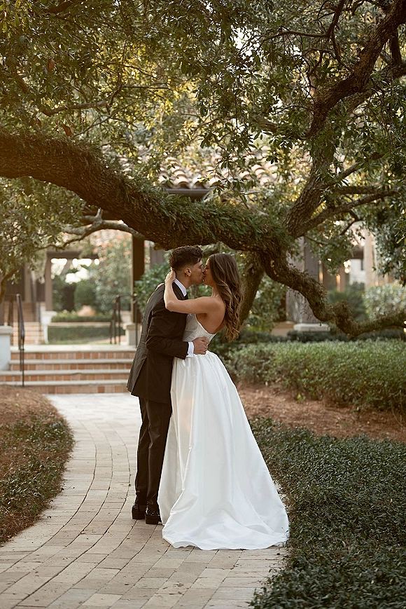 Wedding kiss as bride and groom kissing on a brick walkway in a garden courtyard, her strapless dress and long veil flowing under trees