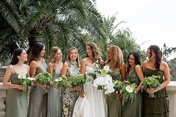 Bridal party portrait with bride and bridesmaids in sage green holding white orchid and anthurium bouquets on a palm-lined terrace