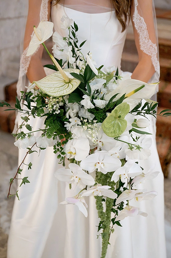 Bridal bouquet of white orchids and anthurium cascading over the bride’s gown on stone steps, with greenery foliage accents