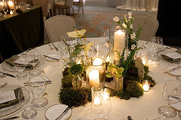 Reception tablescape with wedding centerpiece candles in glass hurricanes, orchids and moss runner, set with chargers, place cards and goblets in a ballroom
