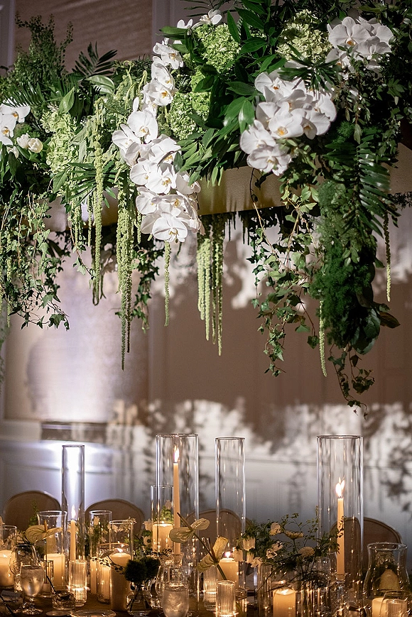 Reception tablescape with wedding table candles in glass cylinder centerpieces, floating candles and orchids beneath overhead greenery indoors
