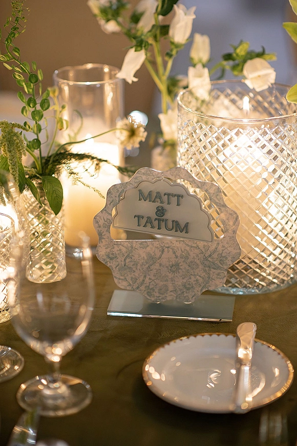 Wedding table decor with a wedding table number sign, glowing glass candles, and white flowers in bud vases on a warmly lit tabletop