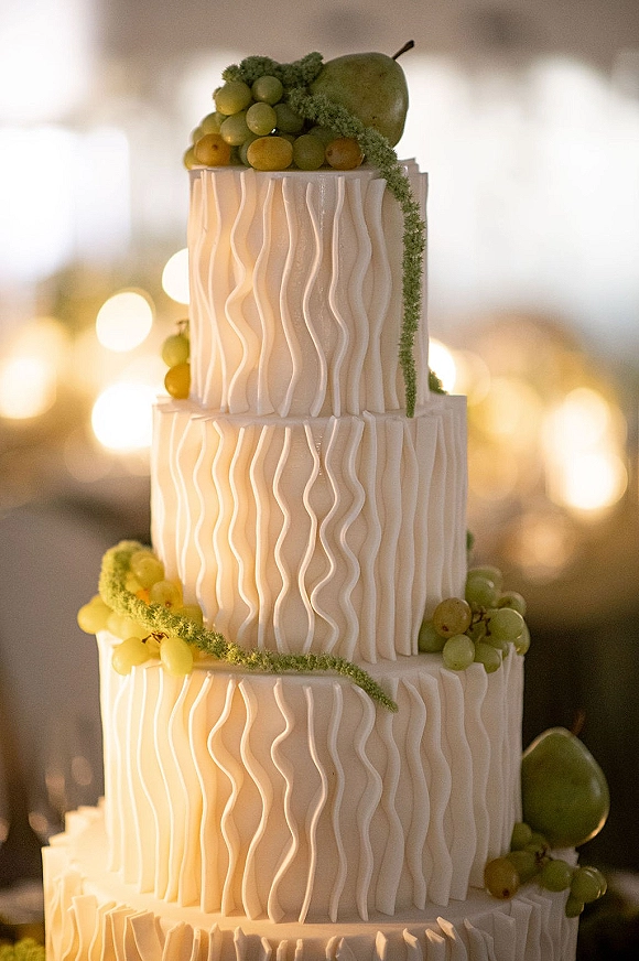Wedding cake with three tier wedding cake design, wavy white buttercream frosting and grape accents, set before soft bokeh lights indoors