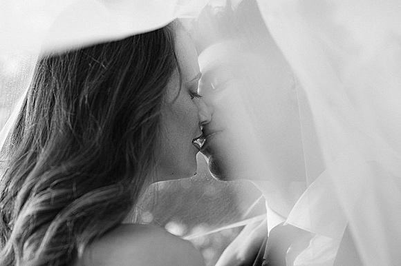 Wedding kiss portrait of bride and groom kiss under a bridal veil, groom in tuxedo with boutonniere in soft blurred outdoor light