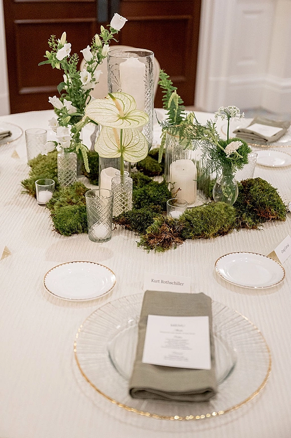 Reception tablescape with moss table runner, white flowers, and pillar candles in glass hurricanes on a white tablecloth indoors