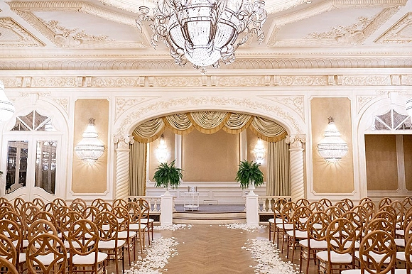 Indoor ceremony setup with a ballroom wedding ceremony aisle lined in white petals, wood chairs, and an arched stage under a crystal chandelier