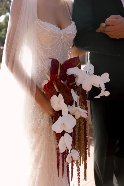 Bridal bouquet with white orchid cascade, burgundy anthuriums and hanging amaranthus held against a beaded dress in sunlit greenery