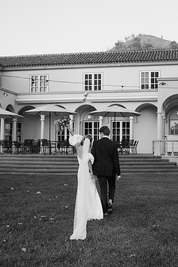 Newlywed couple walking away, bride holding bouquet up, her veil and long train flowing past patio umbrellas and arched facade