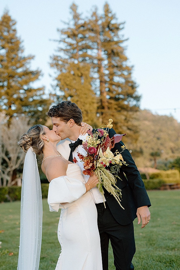 Wedding kiss portrait of bride and groom kissing in a close embrace, bride holding bouquet with veil, lawn and tall trees behind