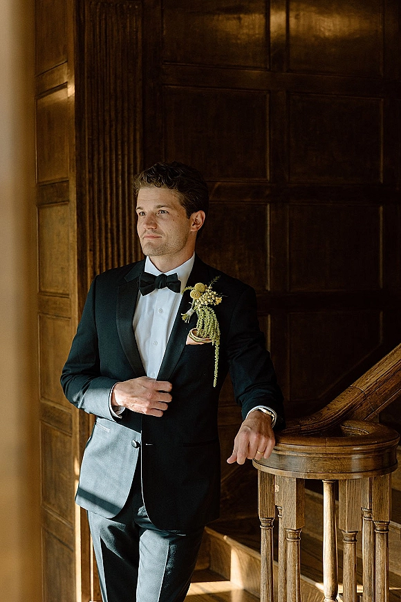 Groom portrait in a black tuxedo, bow tie, and boutonniere, leaning on a wooden banister in a window-lit hallway