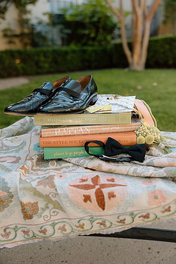 Groom accessories flat lay with black shoes and bow tie on stacked books, paper cards and floral sprig on a lawn with hedges