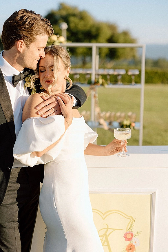 Couple portrait of bride and groom embrace at an outdoor bar counter, bride in strapless dress holding a champagne coupe on a lawn backdrop