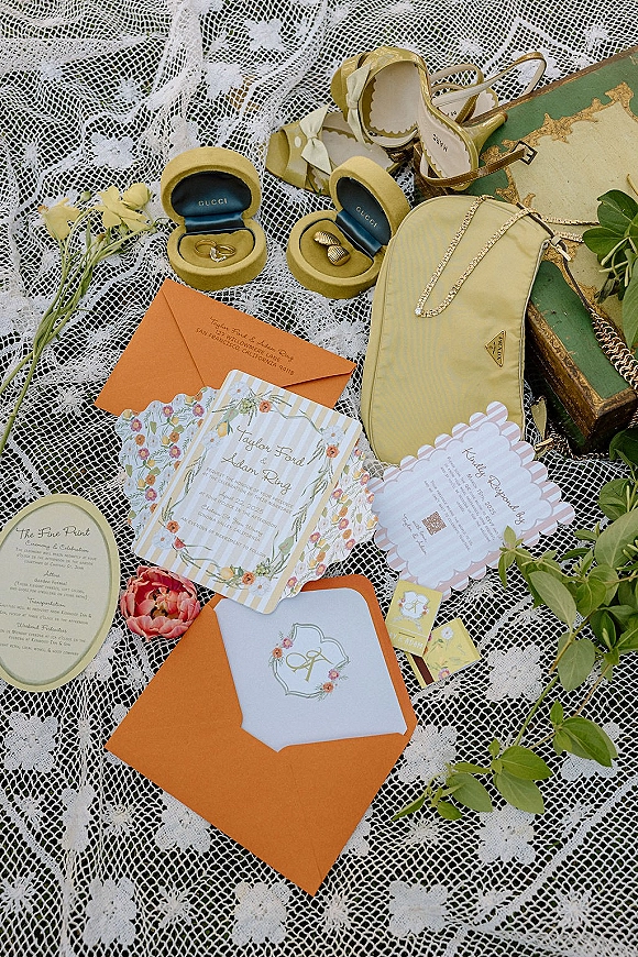 Invitation suite wedding flat lay on lace fabric with floral cards, orange envelopes, wax seal, rings, heels, clutch, and flowers