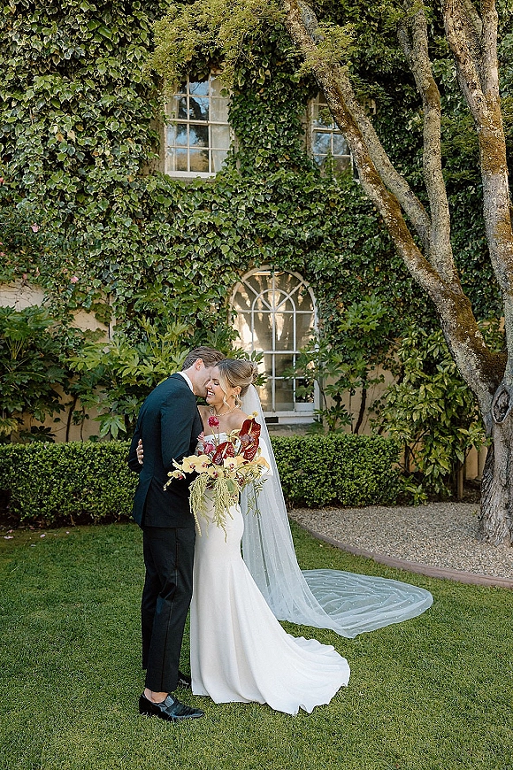 Couple portrait of bride and groom embrace in a forehead kiss, bride’s veil and bouquet flowing beside an ivy wall with arched window